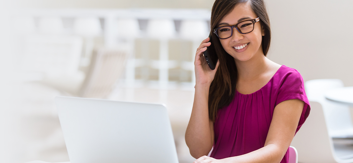 Woman with glasses, sitting in front of a desktop, while on the phone.